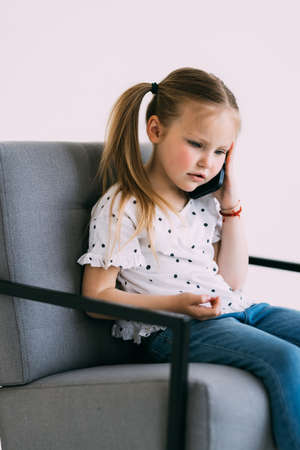 Young little confused girl talk phone sit on chair on white backgroundの写真素材