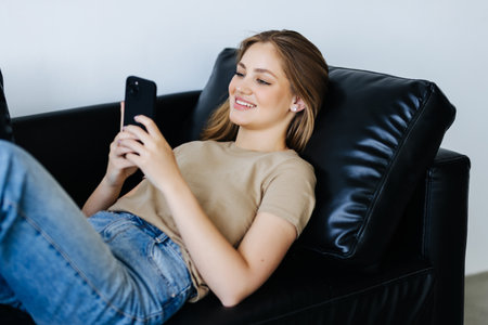 Young woman sit relax on sofa in living room browsing surfing wireless internet on smartphone,...