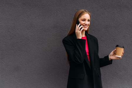 happy woman with coffee to go looking at camera while talking on smartphone outdoors
