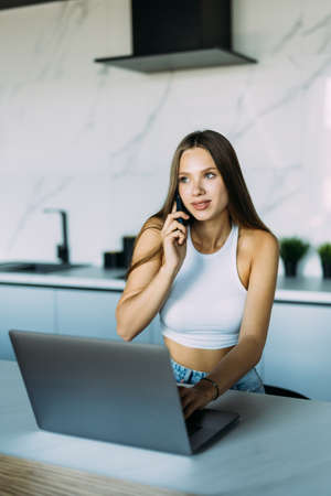 Photo of a young cheerful happy woman indoors at home at the kitchen using laptop computer talking by mobile phone.の写真素材