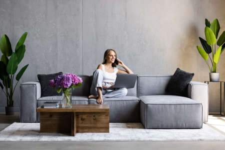 Cute smiling woman using laptop and talking on cellphone while sitting on sofa at living roomの写真素材
