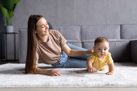 Happy young mother watching her cute baby crawl on floor at homeの写真素材