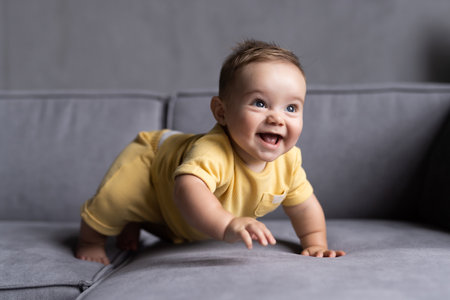 Portrait of lovely laughing baby boy with blue eyes and blue shirt sitting on couch and looking up, blurred toys in background.の写真素材
