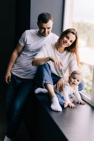 Happy family sitting on windowsill with daughter. Family smiling while sitting together in front of...