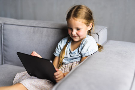 Smiling little girl lying on comfortable couch, enjoying playing online game on digital tablet computer. Addicted to technology, happy small kid using funny applications, web surfing information.の写真素材
