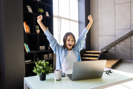 Happy excited woman at home workstation triumphing with raised handsの写真素材