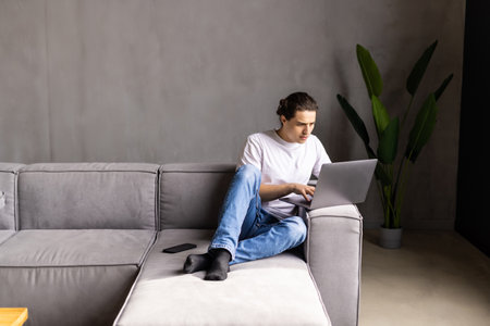 Portrait of young man smiling and using laptop while sitting on sofa at homeの写真素材