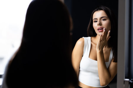 beautiful smiling young woman touching face and looking at mirror in bathroomの写真素材