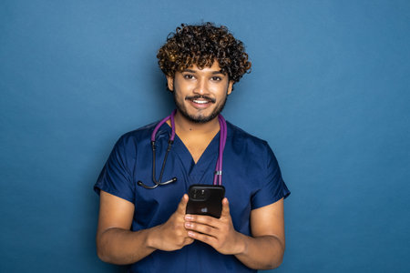 Portrait of pretty indian doctor with stethoscope holding his phone while standind isolated on blue background.の写真素材