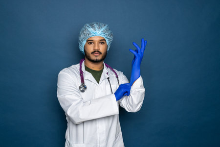 Indian doctor or physician male looking at hand with focused expression as putting on blue surgical...