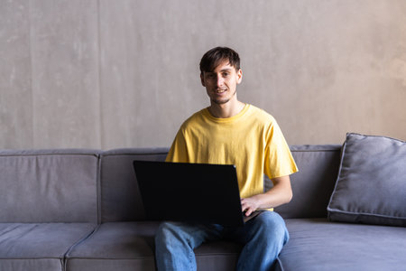 Portrait of young happy bearded man smiling and using laptop while sitting on sofa at homeの写真素材