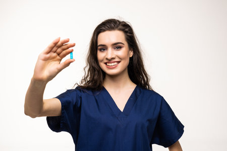 Pointing at pills. Portrait of young woman, doctor, therapeutic or medical advisor wearing face mask and blue uniform on white background. Concept of healthcare and medicineの写真素材