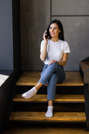 Beautiful woman talking on the phone and working on a laptop on her lap while sitting on the stairs...