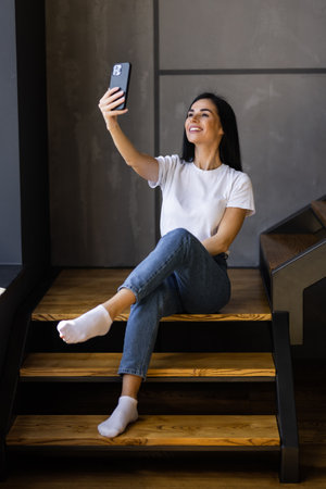 young woman at home making a selfie with smartphone while having a coffee on the stairs of her apartmentの写真素材