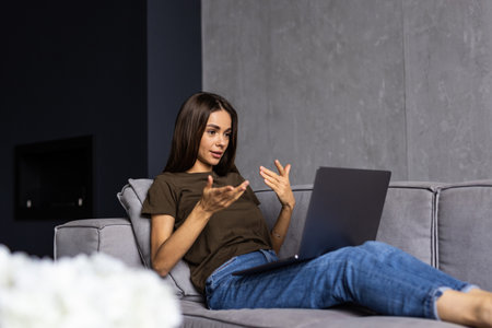 Smiling young woman enjoying morning coffee using laptop sitting at home on sofa, attractive happy girl video calling on computer having fun online or chatting with friends while relaxing on couchの写真素材