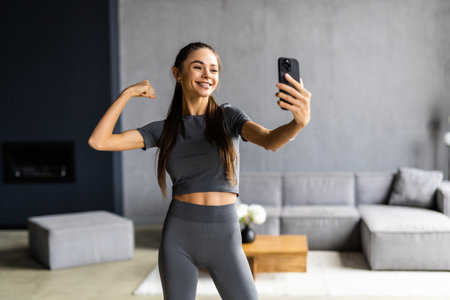 Close up portrait of sportive powerful woman dressed in tight gray top demonstrating her biceps taking selfie or having video call at homeの写真素材