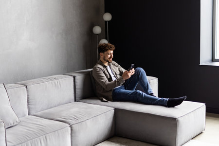 Happy man sitting in living room on couch at home chatting typing email message using cellular,...
