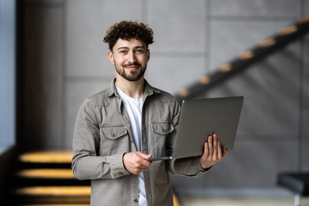 Portrait of handsome mature male office worker with laptop in hands standing in office and smiling...