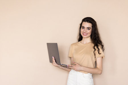 Portrait of gorgeous woman standing and holding laptop isolated over beige backgroundの写真素材