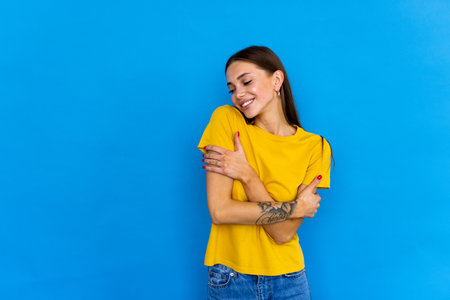 Young woman standing over blue background hugging oneself happy and positive, smiling confident. self love and self careの写真素材