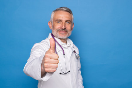 Good-looking cheerful handsome gray-haired middle aged man in medical workwear doctor showing thumb up and smiling at camera, isolated on blue background. Checkup, healthcare, medical insuranceの写真素材