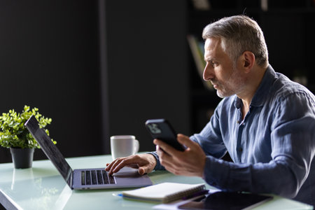Smiling mature businessman holding smartphone sitting in office. Middle aged manager ceo using cell phone mobile apps and laptop. Digital technology applications and solutions for business developmentの写真素材