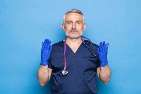 Mature male doctor in medical uniform putting on protective sterile blue gloves while standing against blue background.の写真素材