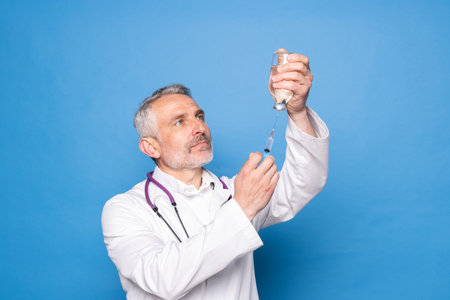 Portrait of joyful medical doctor with stethoscope in gloves holding syringe standing isolated over blue backgroundの写真素材