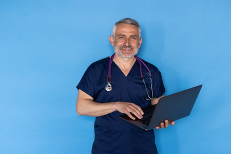 Cheerful handsome middle aged european man wearing medical workwear and eyeglasses doctor holding modern computer laptop over blue studio background, have online appointment with patientの写真素材