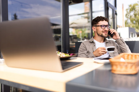 Happy businessman with laptop on table talking over smart phone at outdoor cafeの写真素材
