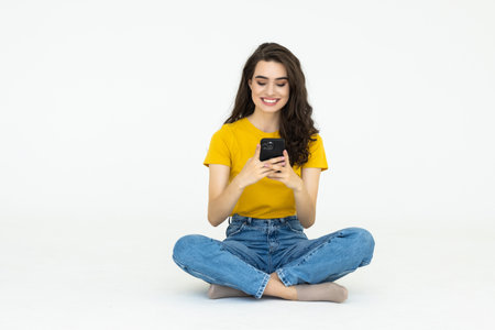 Young mixed race woman sitting on the floor isolated on white background sending a message with the mobileの写真素材