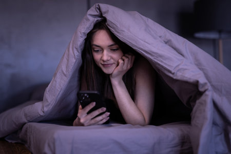 Sleepy woman lying in bed under the blanket using smartphone at nightの写真素材