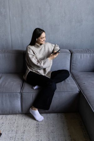 Beautiful brunette woman sitting on a white sofa next to gray and white blanket and pillow, smiling to the camera, holding a phone.の写真素材