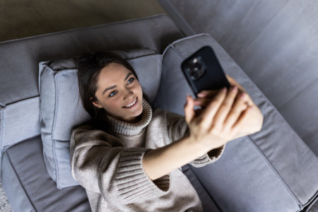 Photo of brunette woman 30s taking selfie on cell phone while sitting on couch in bright apartmentの写真素材