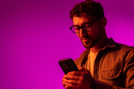 Side view of astonished surprised man in shirt using online service on mobile phone and expressing amazement, looking at camera. Indoor studio shot isolated on colorful neon light background.の写真素材