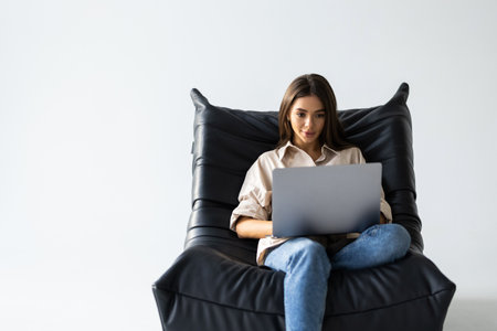 Relaxed young woman sitting in a bag chair with stretched arms using a laptop surrounded by artificial plantsの写真素材
