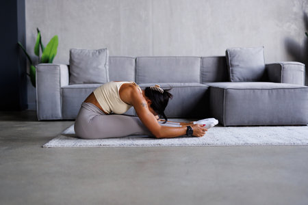 Slim sportswoman in sportswear warming-up muscles before workout doing stretching exercise sitting on floor at homeの写真素材
