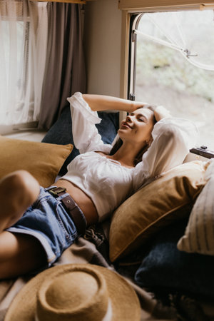 Happy woman sitting on bed inside camper van bedroom and looking outside the window with dreamer expression. Freedom and independence lifeの写真素材