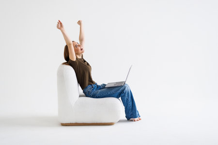 Ecstatic woman sitting in armchair with laptop, lifting hands up, making YES gesture, celebrating online win or success against white wall, free space. Achievement conceptの写真素材