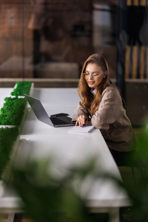 Woman working on a laptop in a modern office with greenery and coffeeの写真素材