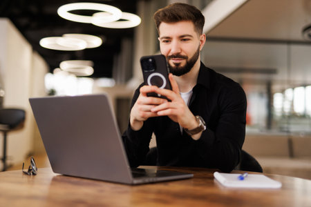 A smiling young male office worker sits in the office at a desk and uses a mobile phone, writes a message, corresponds.の写真素材