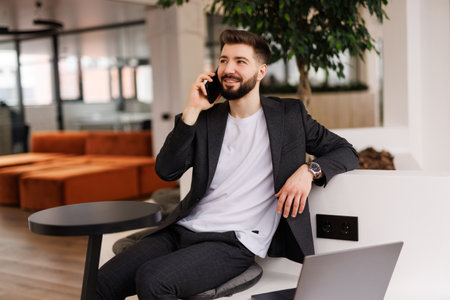 Good business talk. Handsome young man talking on smart phone and using computer while working in the officeの写真素材