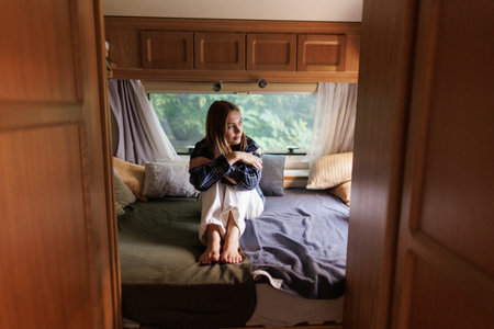 Happy woman sitting on bed inside camper van bedroom and looking outside the window with dreamer expression. Freedom and independence lifeの写真素材