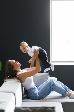 young mother holds and lifts her little son on the sofa, woman plays with her child at home,