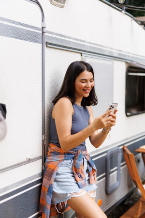 Happy woman standing near camper van, using smartphone on camping trip in countryside, copy space. Cool young lady browsing web, chatting online, watching video on summer vacation outdoorsの写真素材