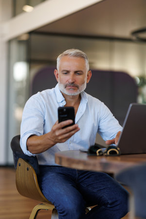 Smiling mature businessman holding smartphone sitting in office. Middle aged manager ceo using cell phone mobile apps and laptop. Digital technology applications and solutions for business developmentの写真素材