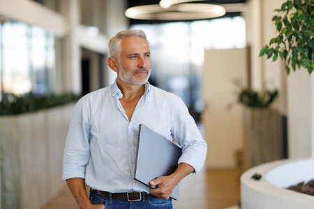 Mature adult man holding laptop in modern office lobby, showing polished floor and greenery. Professional, corporate, workspace, technology, executive, leadership, minimalistの写真素材