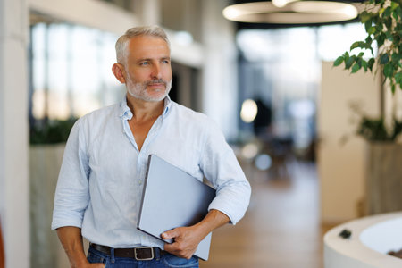 Happy middle aged man wearing suit looking at camera holding laptop standing in officeの写真素材