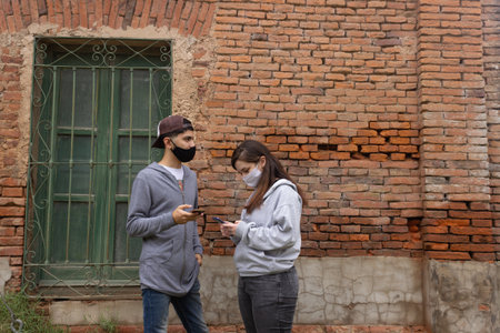 Asian woman and man wearing face mask using mobile phone in front of brick wall