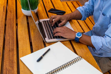 Hands typing a laptop on a desk, next to a cell phone, a notebook and a pen